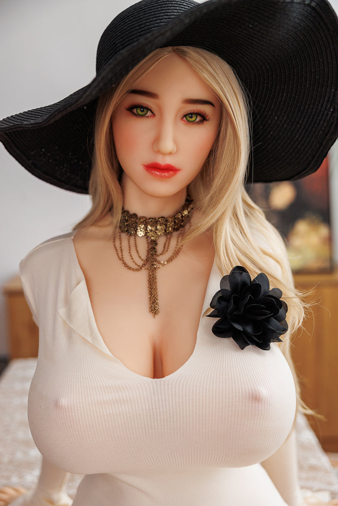 Della, realistic full-body TPE sex doll with long blonde hair and green eyes, dressed in a fitted white off-the-shoulder top, enhanced with a large black flower and layered gold necklace, wearing a wide-brimmed black sun hat, set in a cozy indoor backdrop.