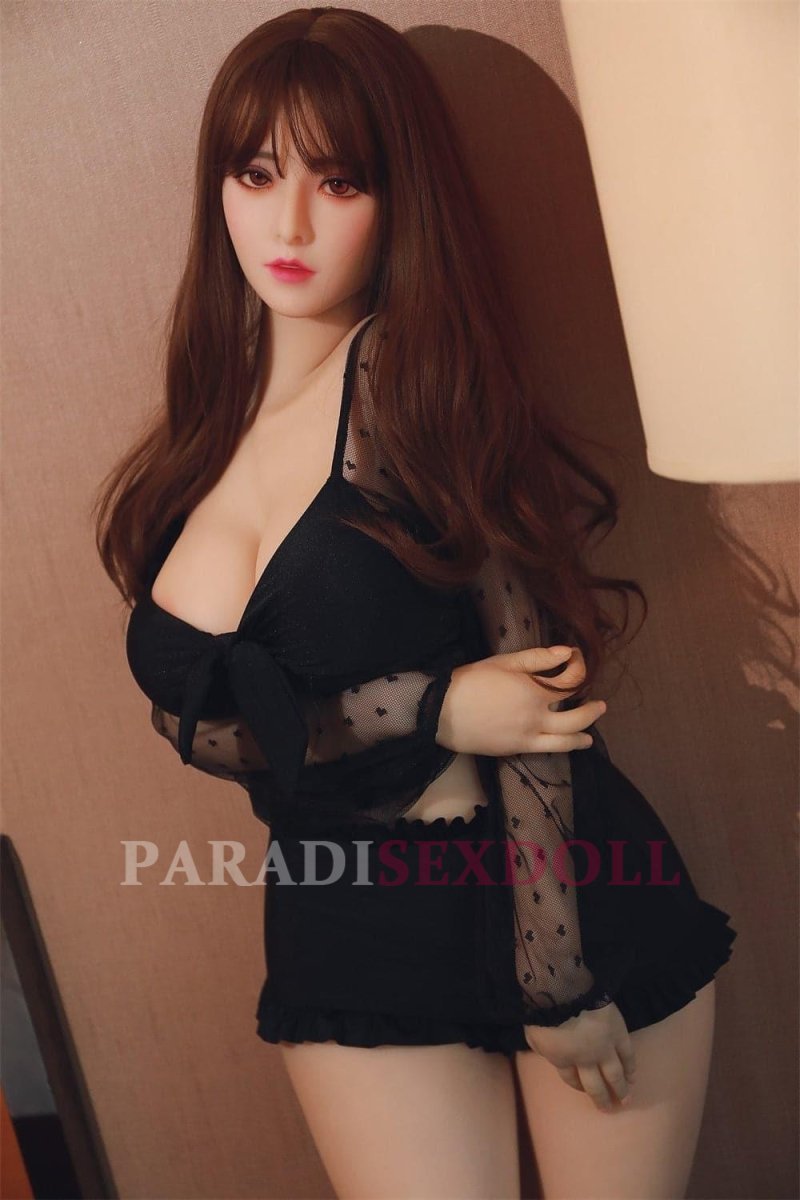 Korean life-size sex doll Natalie posed elegantly against a neutral backdrop, wearing a black sheer polka-dot top and high-waisted shorts, showcasing her long hair and realistic skin texture in a softly lit environment.