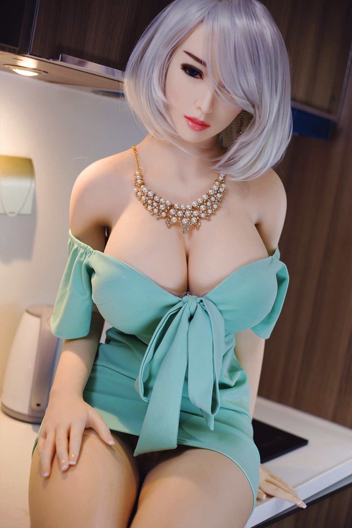 170cm Akiko realistic sex doll wearing a fitted light green off-shoulder dress with a bow and a pearl necklace, highlighting her K-Cup breasts and styled short platinum blonde hair, posed elegantly in a modern kitchen setting.