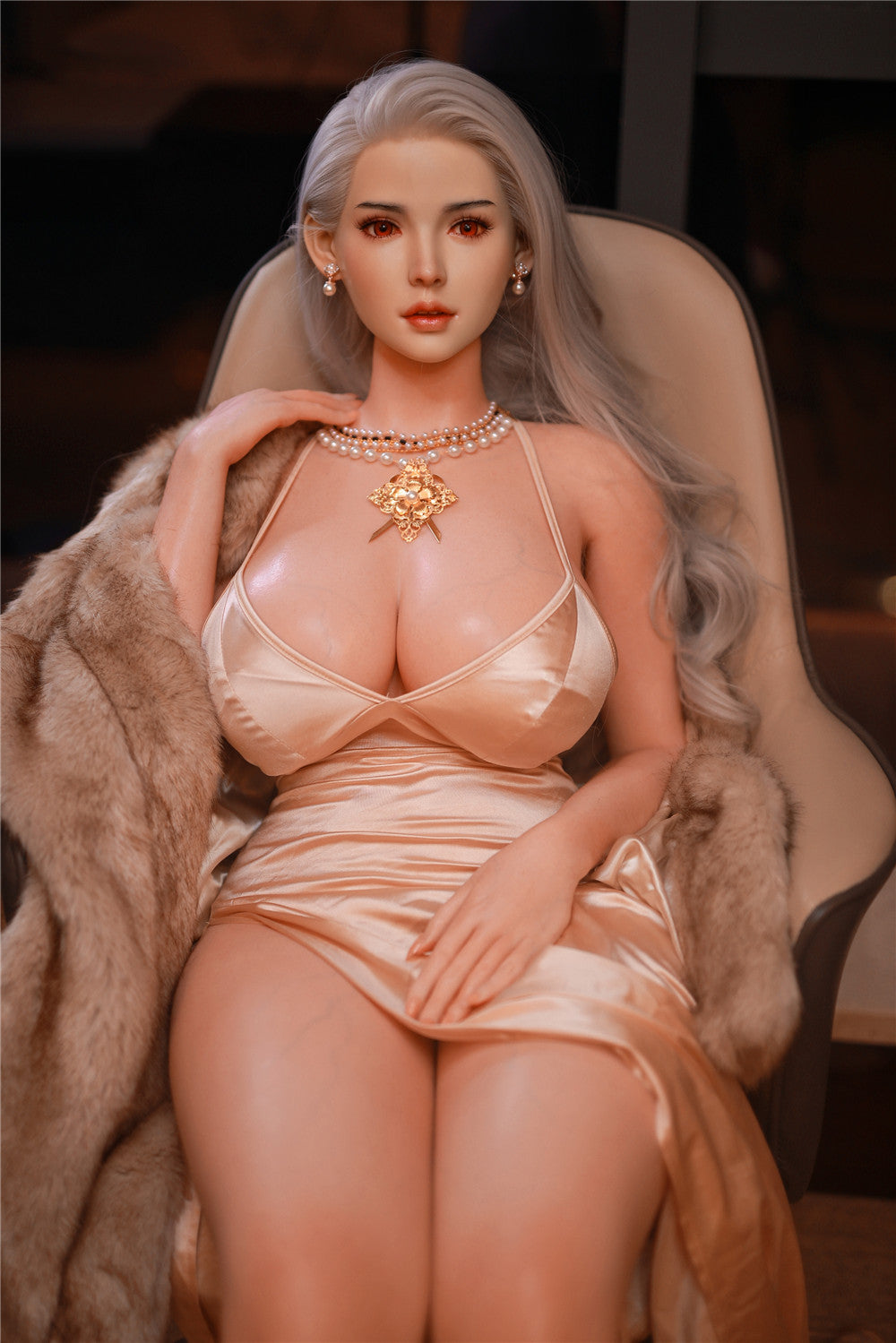 162cm Cathy doll styled in a luxurious champagne satin gown with a thigh-high slit, adorned with an intricate gold necklace and a glamorous faux fur coat, gracefully seated in a modern chair with warm ambient lighting.