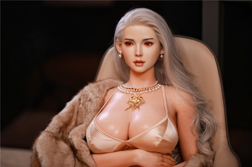 Elegant 162cm Cathy doll in a glamorous satin top with a deep neckline, adorned with an intricate gold necklace featuring a floral accent. She is elegantly seated in a modern chair, draped with a luxurious fur coat under warm ambient lighting, showcasing her lifelike features.