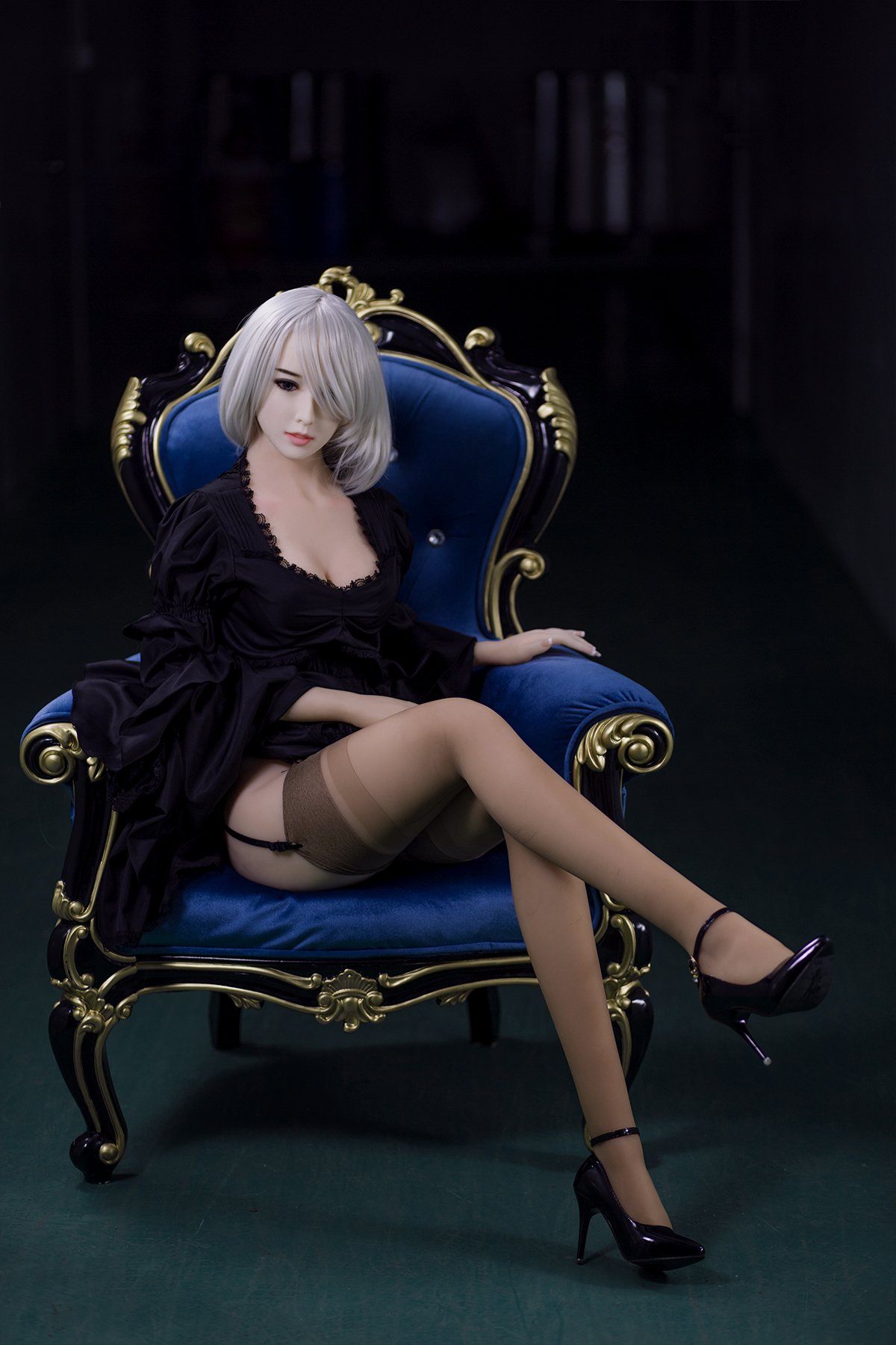 170cm Akiko realistic sex doll elegantly seated on a royal blue armchair, showcasing her K-Cup breasts in a fitted black lace robe and sheer stockings. The doll displays a chic short platinum blonde hairstyle and high-heel shoes, posed seductively in a darker setting.