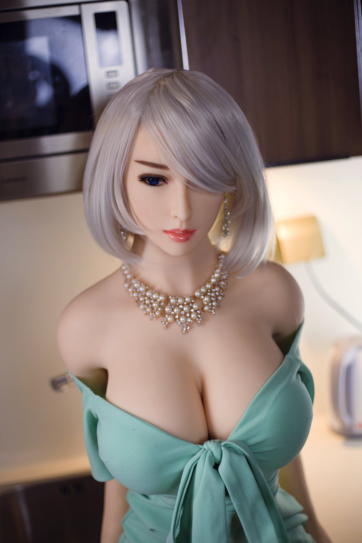 Akiko 170cm realistic sex doll in a light green off-shoulder dress with a bow, showcasing K-Cup breasts and a pearl necklace, posed elegantly in a modern kitchen environment with contemporary appliances.