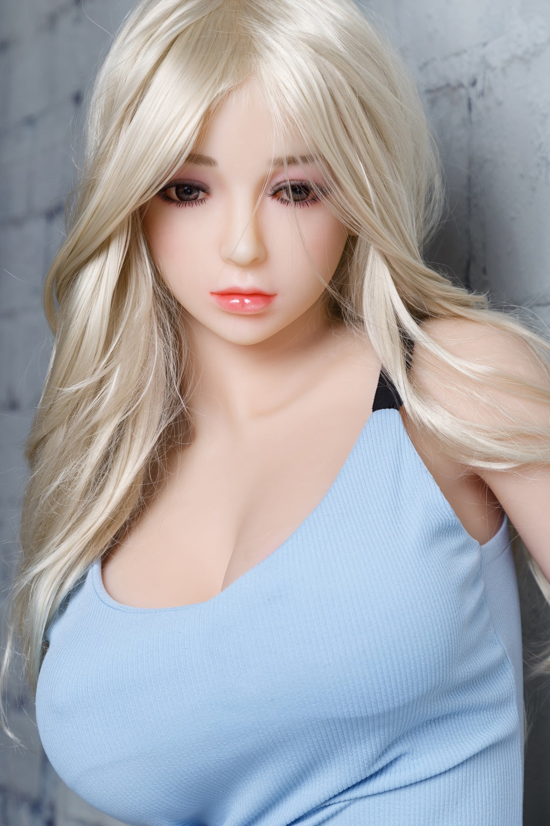 Gloria, a 5ft 5 realistic sex doll with long blonde hair, wearing a fitted light blue tank top that accentuates her large breasts, posed against a textured gray brick background, emphasizing her lifelike features and inviting expression. Ideal for intimate companionship and adult enjoyment.