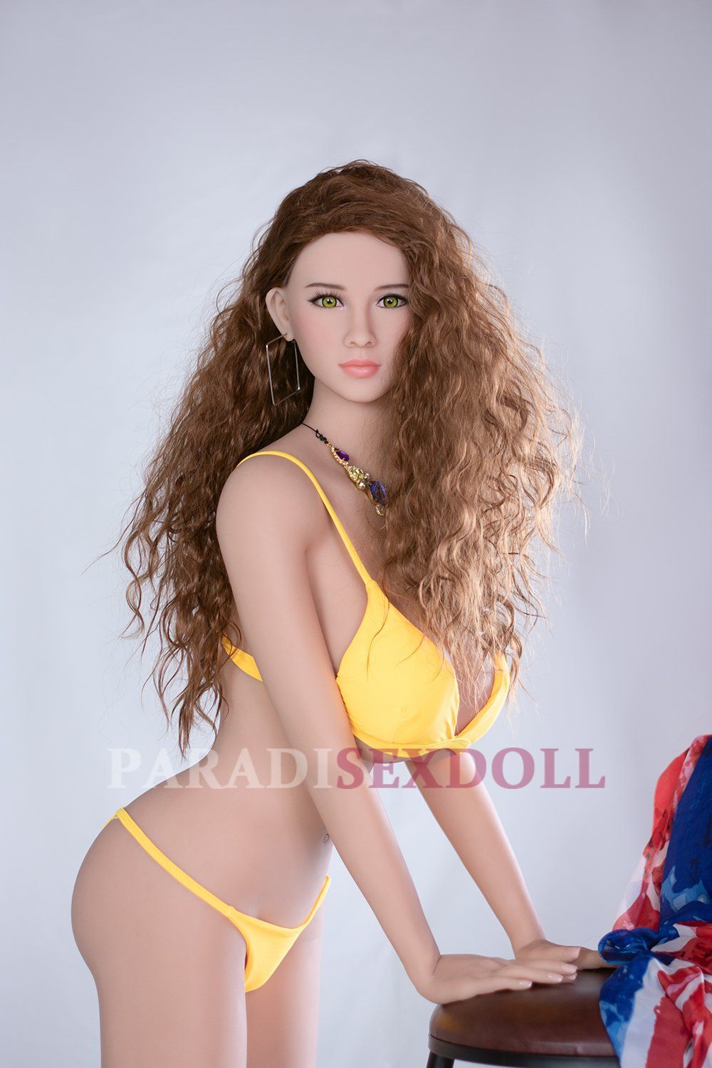 Lifelike K-Cup sex doll with long, wavy brown hair and bright green eyes, posing elegantly on a bar stool in a vibrant yellow bikini, complemented by a colorful necklace and shawl.