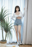 Full-size realistic silicone sex doll in white lingerie and denim shorts standing by window