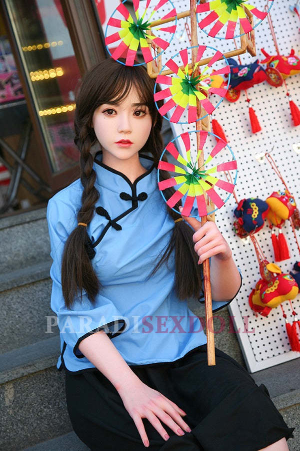 Lifelike Asian silicone sex doll with long braids in a blue traditional Chinese shirt, seated with colorful decorative wheels, in front of a background of traditional Chinese crafts.