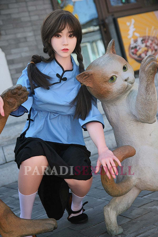 Realistic Asian silicone sex doll in a blue traditional Chinese shirt and black skirt, kneeling next to a playful cat statue in an outdoor urban setting, showcasing her braided hair and vibrant attire.