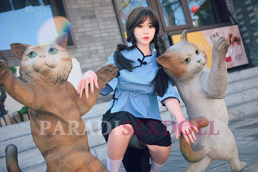 Lifelike Asian silicone sex doll with long braided hair wearing a light blue traditional Chinese shirt and black skirt, crouching playfully between two whimsical cat statues in an urban outdoor setting.