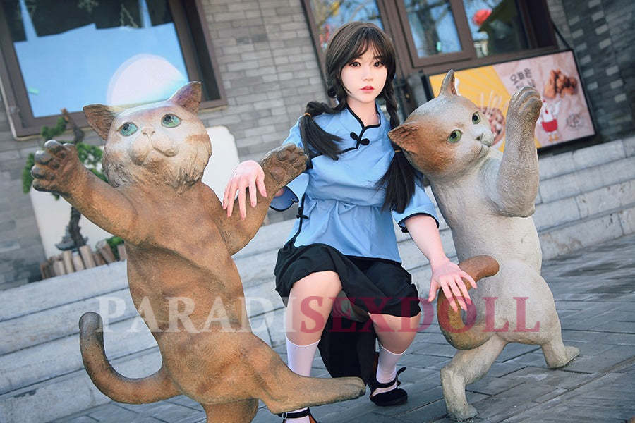 Realistic Asian silicone sex doll with long braided hair, dressed in a blue traditional Chinese shirt and black skirt, crouching playfully between two whimsical cat statues in an urban outdoor setting.