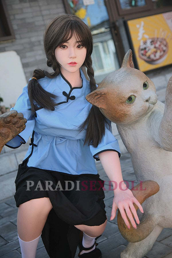 Realistic Asian silicone sex doll wearing a blue Chinese shirt and black skirt, posed next to a playful cat statue in an outdoor urban setting, with her braided hair styled neatly.