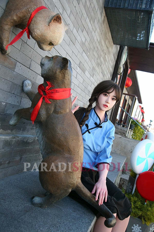 Realistic Asian silicone sex doll with long braided hair, dressed in a blue traditional Chinese shirt and black skirt, seated next to a playful cat statue wearing a red ribbon, with a textured stone wall background.