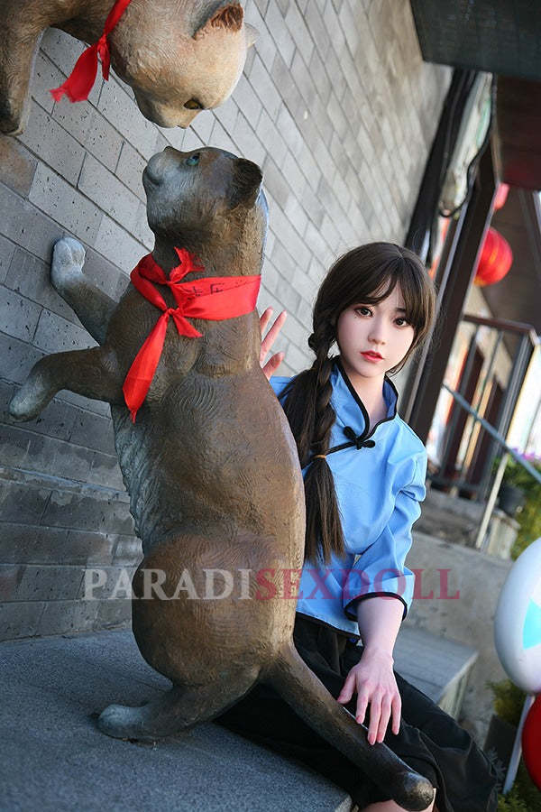 Realistic Asian silicone sex doll with long braided hair in a blue traditional Chinese shirt and black pants, seated next to a whimsical cat statue with a red ribbon, posed against a textured stone wall in an outdoor urban setting.