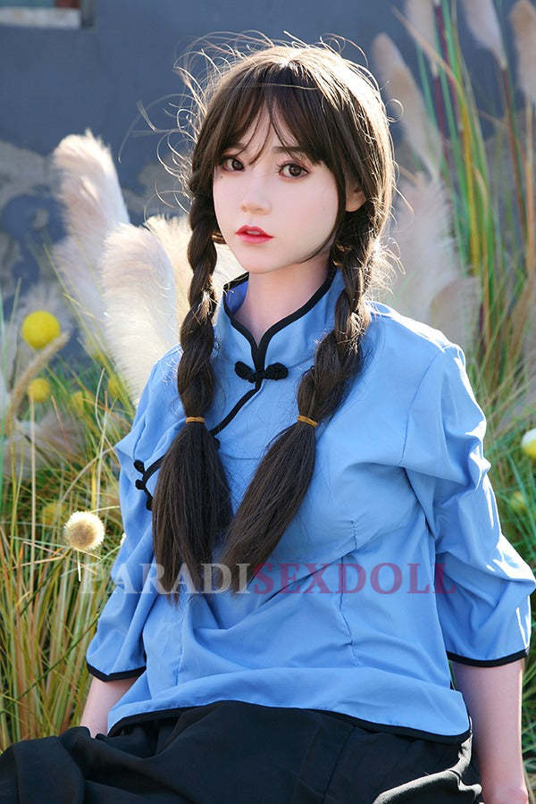 Lifelike Asian silicone sex doll with long braided hair in a blue traditional Chinese shirt, seated in a natural outdoor setting surrounded by tall grass and yellow flowers.