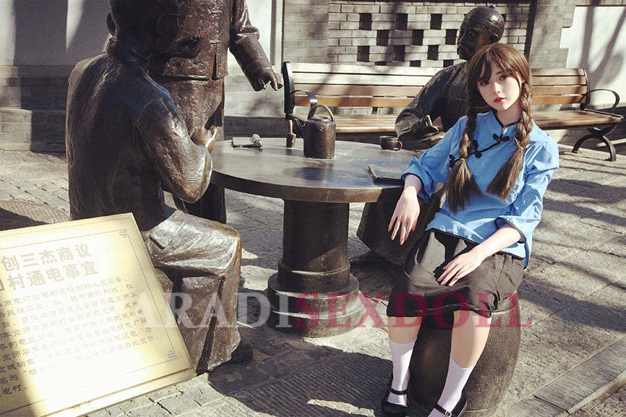 Asian silicone sex doll with long braided hair, dressed in a blue traditional Chinese shirt and black skirt, seated at a round table in an urban outdoor setting, alongside bronze statues of people, with a historical plaque visible nearby.