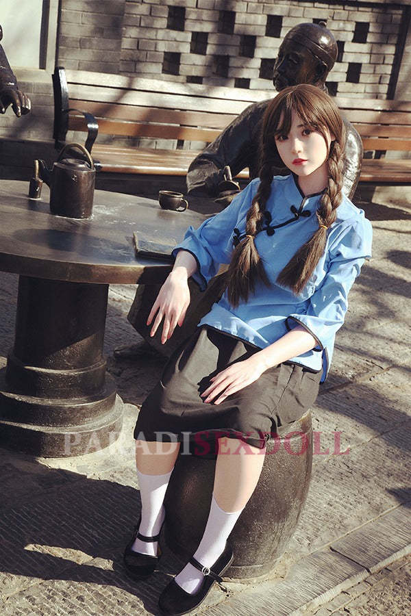 Asian silicone sex doll with long braided hair dressed in a light blue traditional Chinese shirt and black skirt, seated on a stool at an outdoor table, surrounded by a vintage urban backdrop with stone walls and a statue.