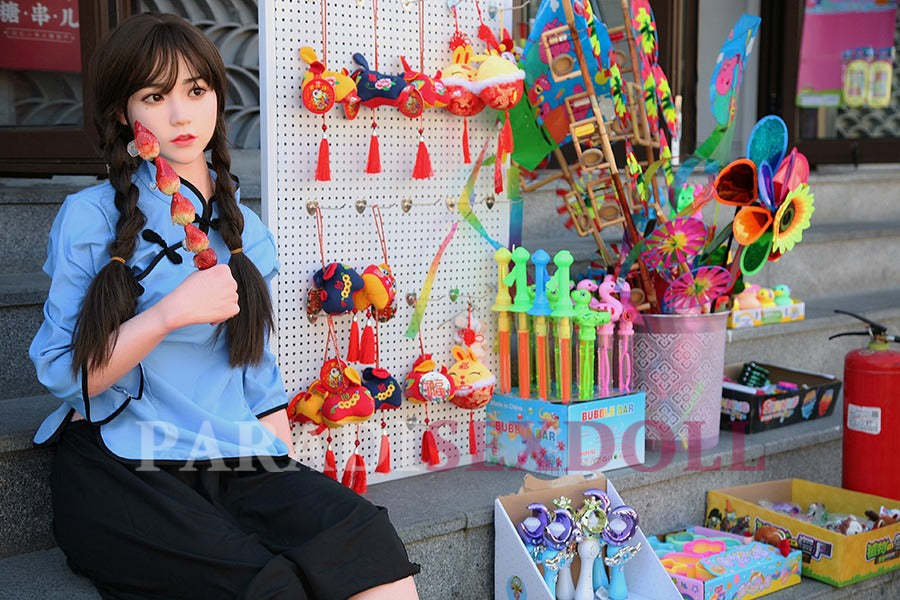 Lifelike Asian silicone sex doll dressed in a blue traditional Chinese shirt and black skirt, seated outside near a colorful display of toys and decorative items, with long braided hair and a playful expression.