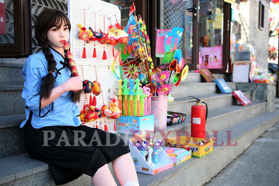 Realistic Asian silicone sex doll in a blue traditional Chinese shirt and black pants, seated on outdoor steps, playing a flute and surrounded by colorful toys and decorations, against a backdrop of a vibrant street shop.