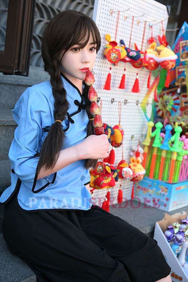 Asian silicone sex doll in a blue traditional Chinese outfit, seated on steps and holding a colorful fruit skewer, surrounded by vibrant Chinese crafts and toys.