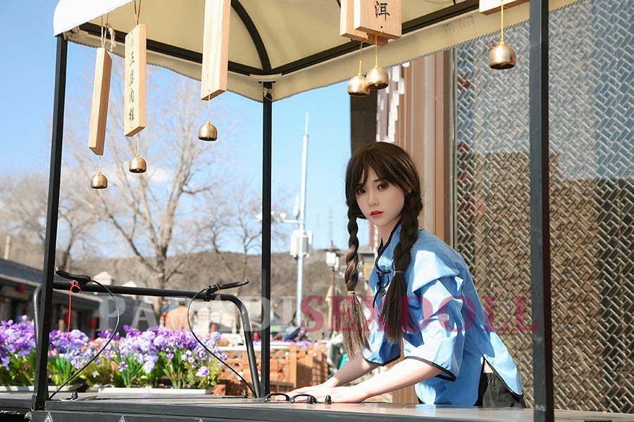 Lifelike Asian silicone sex doll with long braided hair in a blue traditional Chinese shirt, seated at a decorative cart surrounded by vibrant purple flowers and a textured backdrop in an outdoor market setting.