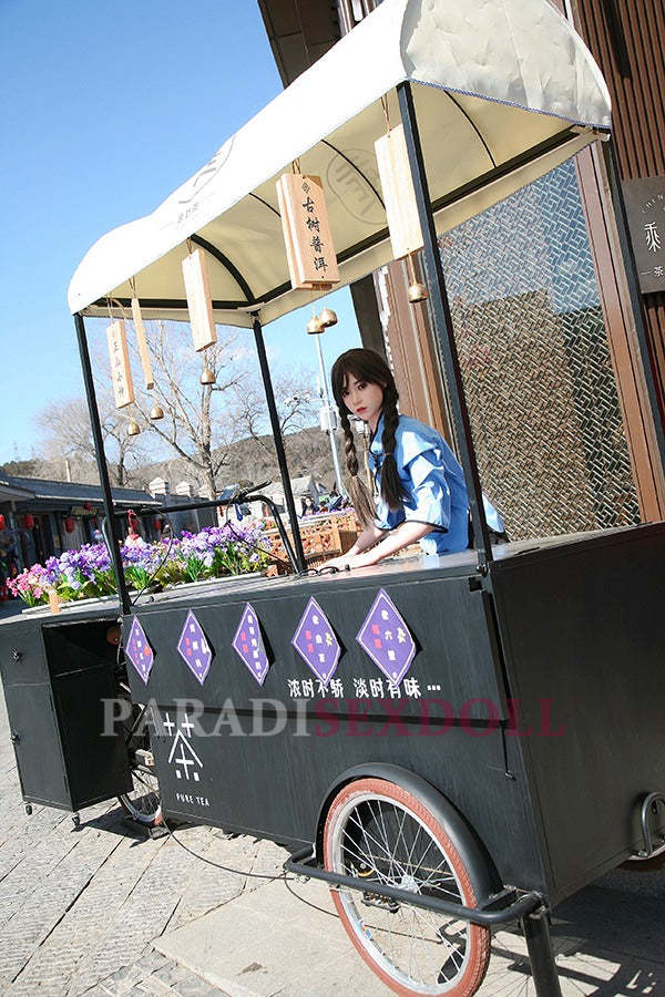 Asian silicone sex doll with long braided hair wearing a blue traditional Chinese shirt, positioned at a street tea cart, surrounded by colorful flowers and decorative tea signage.