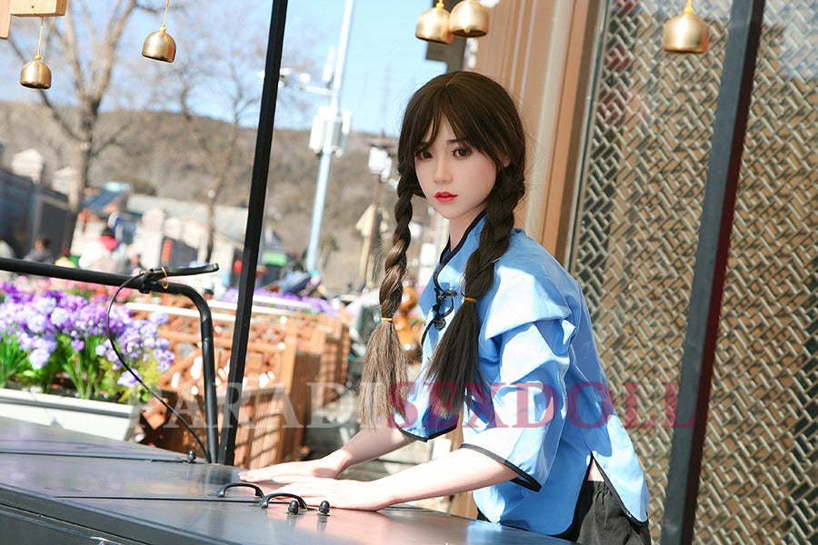 Lifelike Asian silicone sex doll with long braided hair wearing a blue traditional Chinese shirt, standing at a cart in an outdoor setting with vibrant flowers and ornamental decorations in the background.