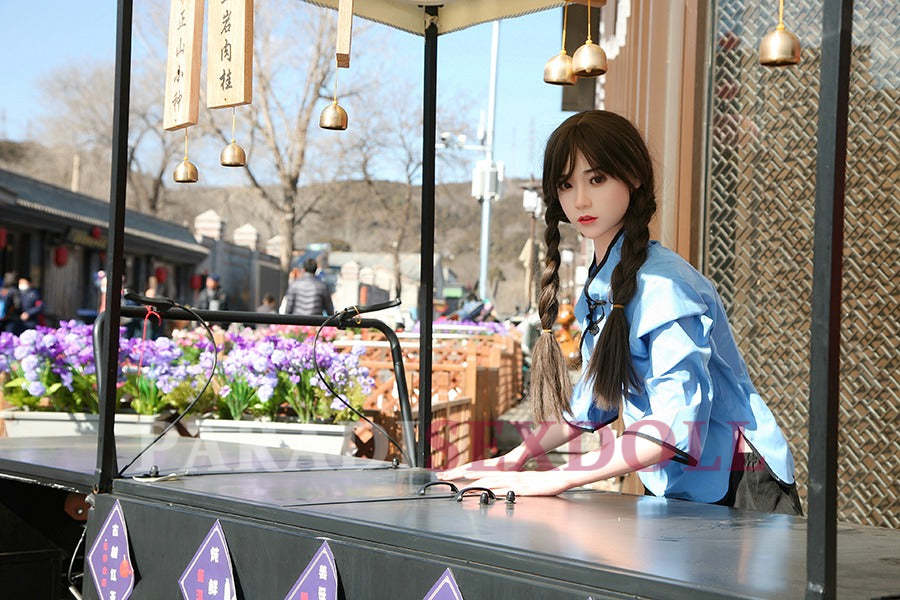 Lifelike Asian silicone sex doll in a blue traditional Chinese shirt and black pants, posed at an outdoor cart decorated with colorful flowers, set against a bustling urban market backdrop.