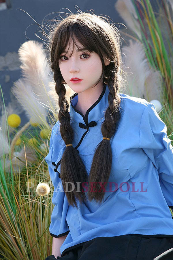 Realistic Asian silicone sex doll with long braided hair, dressed in a blue traditional Chinese shirt, gracefully posed outdoors amidst soft grasses and decorative plants.