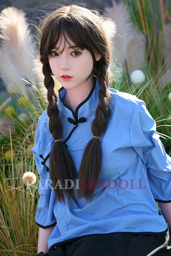 Realistic Asian silicone sex doll with long braided hair, dressed in a blue traditional Chinese shirt with a mandarin collar, posed outdoors amongst tall grasses, showcasing her lifelike features and vibrant attire.