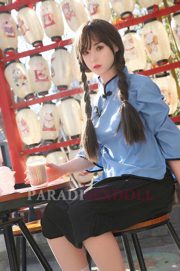 Realistic Asian silicone sex doll with long braided hair, wearing a blue traditional Chinese shirt and black skirt, sitting at a table with a drink, surrounded by decorative Chinese lanterns in a vibrant outdoor setting.