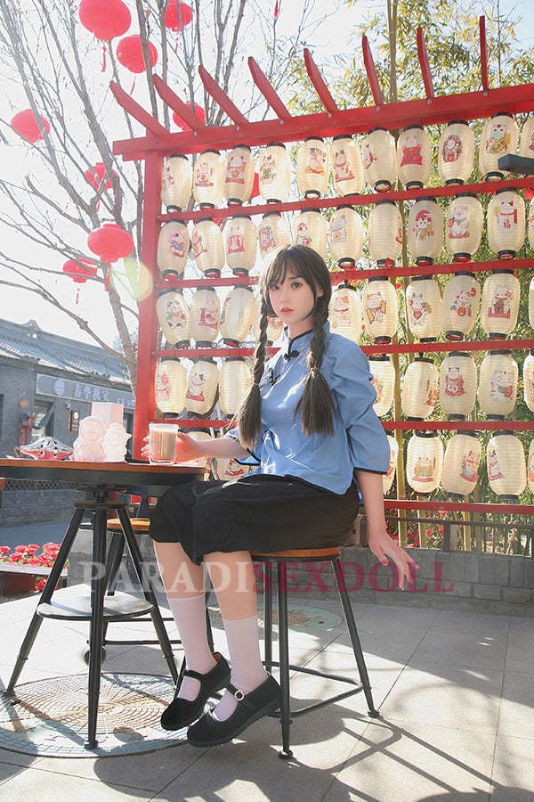 Realistic Asian silicone sex doll wearing a blue traditional Chinese shirt and black skirt, seated at a table outdoors, holding a drink, with decorative Chinese lanterns and vibrant red accents in the background.