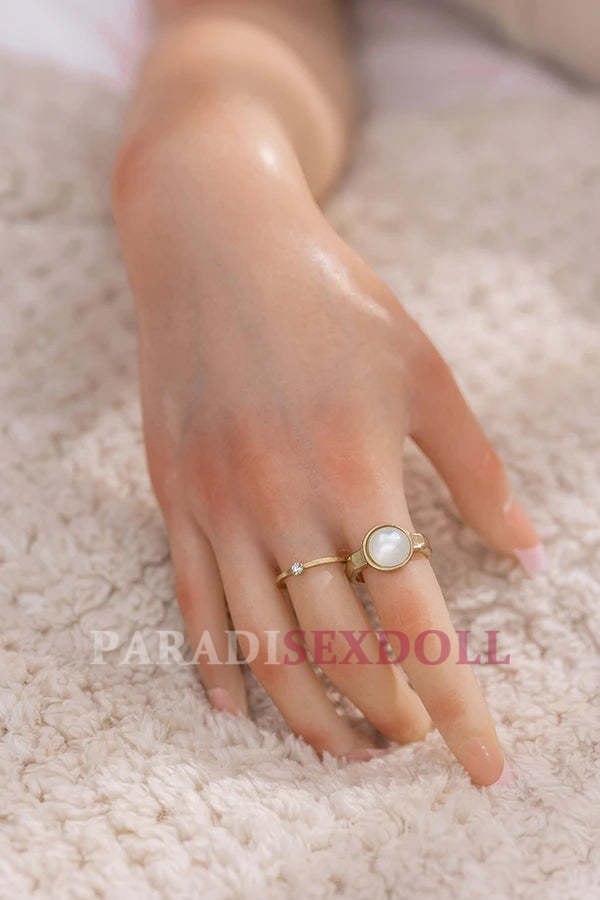 Close-up of the delicate hand of Asian lifelike full silicone sex doll Charlene adorned with a gold ring featuring a pearl stone, resting on a soft, textured beige blanket, showcasing the realistic skin texture and fine details.