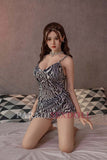 Asian silicone sex doll Qiuyue, 170cm, dressed in a fashionable zebra-print outfit, kneeling on a bed with a modern geometric-patterned pillow, featuring realistic facial and body details.