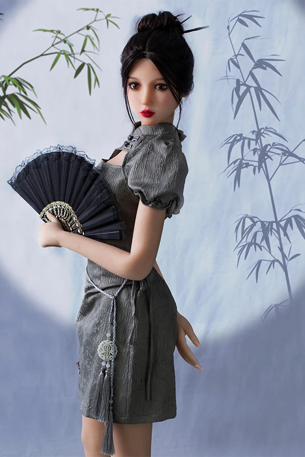 Graceful lifesize Chinese sex doll Winni with small breasts, elegantly posed in a chic gray puff-sleeve dress, holding a black fan against a tranquil bamboo-patterned backdrop.