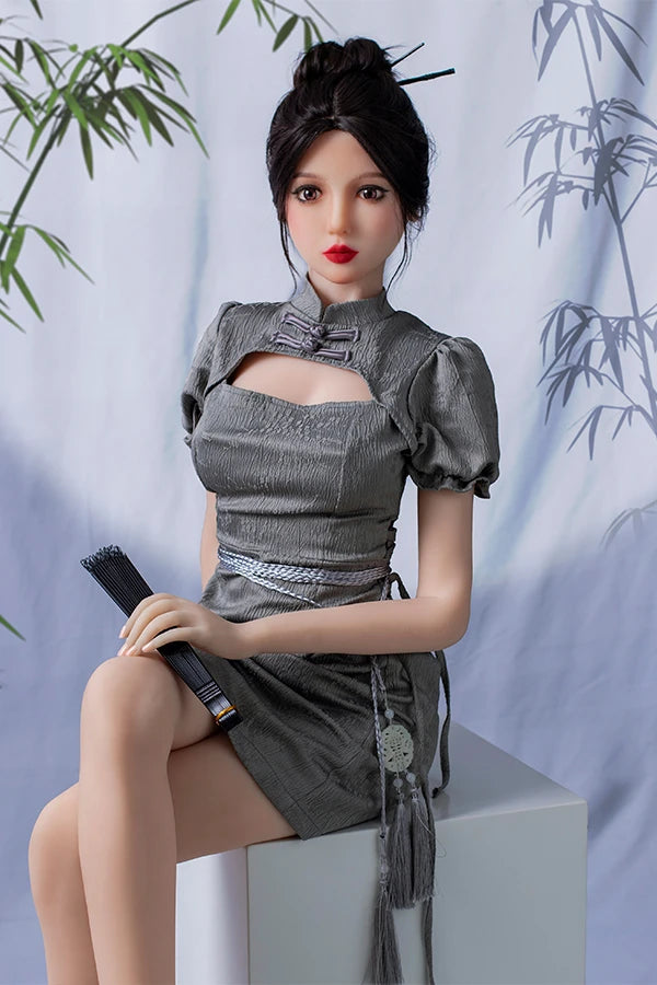 Lifesize Chinese sex doll Winni with small breasts sitting on a white pedestal, wearing a silver dress with puff sleeves, holding a black fan, against a bamboo-patterned background.