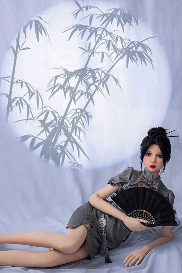 Elegant lifesize Chinese sex doll Winni reclining gracefully while holding a black lace fan in a stylish gray puff-sleeve dress, against a soft backdrop featuring a bamboo shadow.