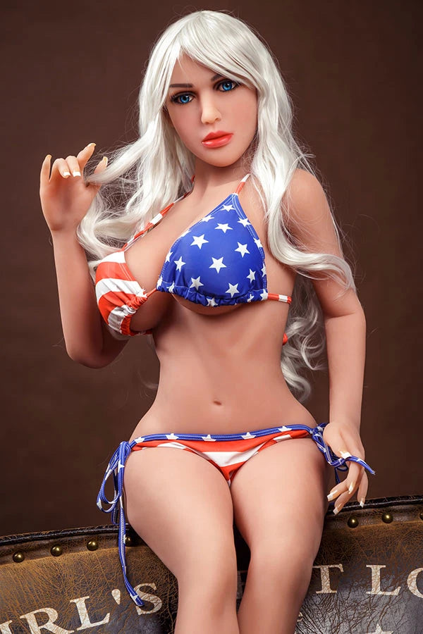 American Beauty love doll Avianna showcasing patriotic red, white, and blue bikini with stars and stripes, featuring long flowing blonde hair and striking blue eyes, playfully posed with one hand touching her hair.