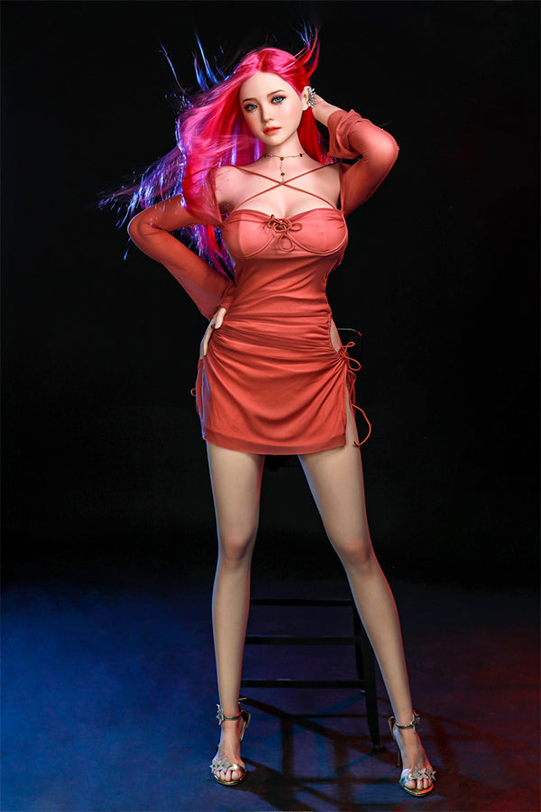 Rana silicone sex doll with vibrant long pink hair and bright blue eyes, elegantly standing in a fitted coral dress, highlighting her lifelike details and inviting expression against a dramatic dark backdrop.