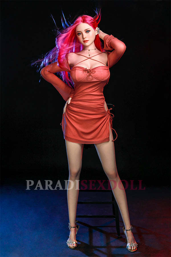 Rana silicone sex doll with vibrant long pink hair and bright blue eyes, elegantly standing in a fitted coral dress, highlighting her lifelike details and inviting expression against a dramatic dark backdrop.