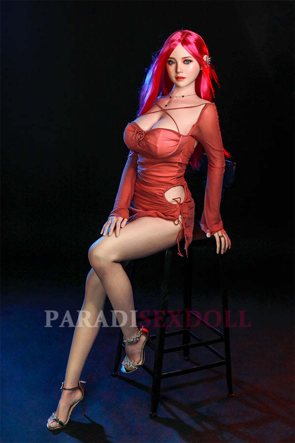 Rana silicone sex doll with long bright pink hair and captivating blue eyes, elegantly seated on a high stool in a fitted coral dress, showcasing her lifelike details and inviting expression in a studio setting.