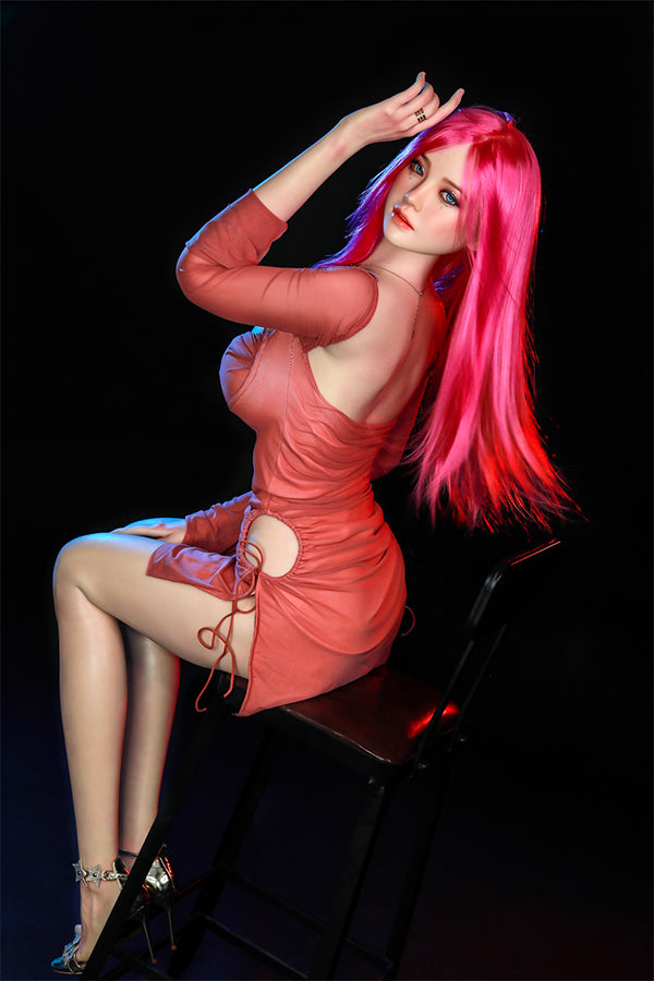 Lifelike Rana silicone sex doll with long bright pink hair and blue eyes, elegantly posed on a chair in a fitted coral dress, showcasing her realistic features and inviting expression.