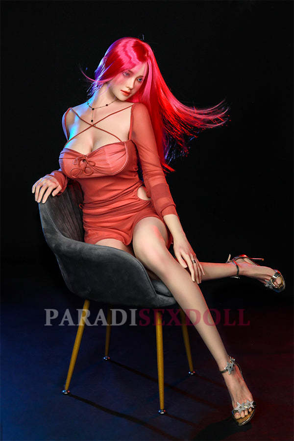 Hyper-realistic Rana silicone sex doll with long bright pink hair and blue eyes, seated gracefully in a stylish chair, wearing a fitted coral dress and showing lifelike features, enhancing her allure and captivating appearance.